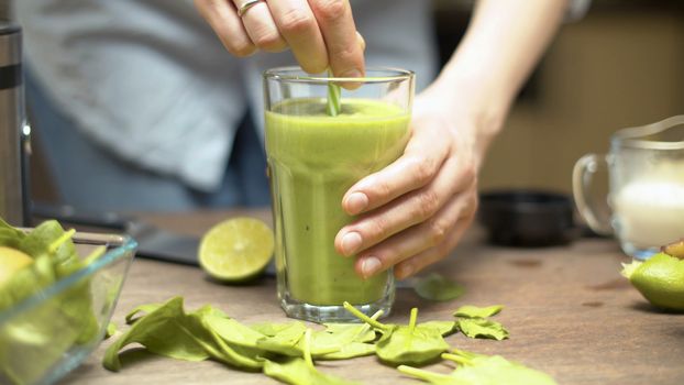 Close up glass of green smoothies on the table. Female hands mixing it with drinking straw. Healthy lifestyle and eating concept