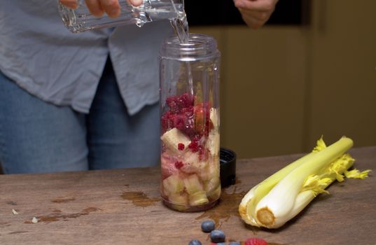 Close up woman's hands pouring water in blender glass with banana, raspberries and celery slices. Healthy eating concept