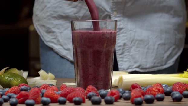 Close up woman's hands pouring ready made smoothies into the glass. Celery, raspberries, blueberries on the table closeup. Fresh healthy food
