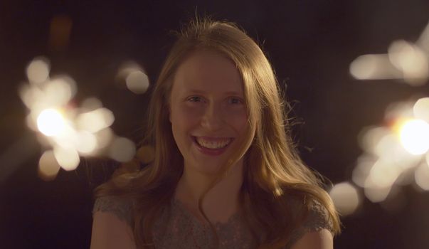 Close up portrait of beautiful caucasian woman celebrating indoors. Blurred sparklers in the foreground. Blond smiling lady on the party looking at camera