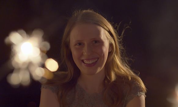 Close up portrait of beautiful caucasian woman celebrating indoors. Blurred sparklers in the foreground. Blond smiling lady on the party looking at camera