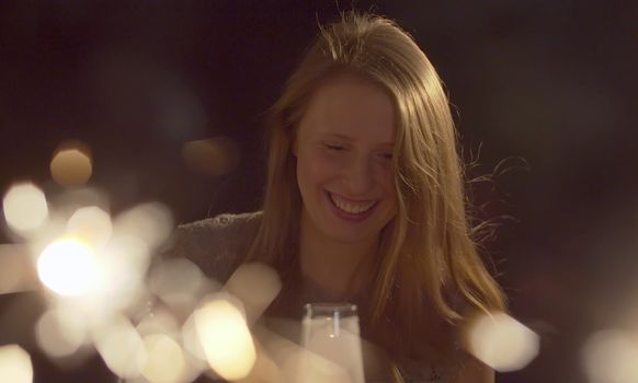 Close up portrait of beautiful young woman drinking champagne on the party. Blurred sparklers in the foreground. Blond smiling woman celebrating indoors. Front view