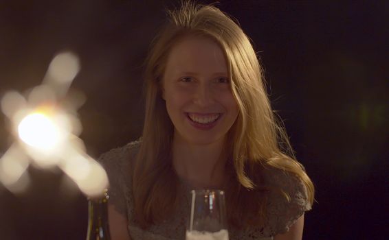 Close up portrait of beautiful young woman drinking champagne on the party. Blurred sparklers in the foreground. Blond smiling woman celebrating indoors. Front view