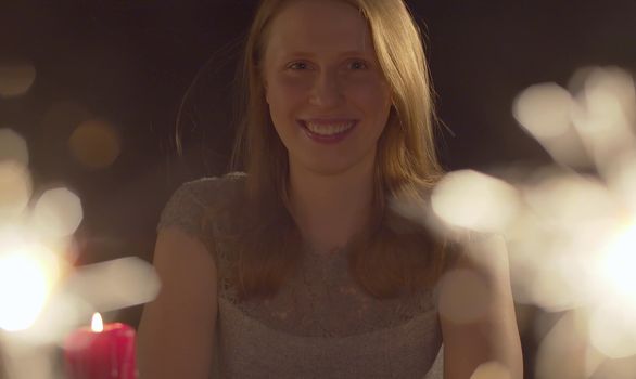 Close up portrait of beautiful caucasian woman celebrating indoors. Burning candle and blurred sparklers in the foreground. Blond laughing girl on the party looking at camera