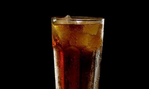 Close up full misted glass of cold cola with ice on black background. Bubbles on ice and water droplets on the glass. Closeup fizzy drink