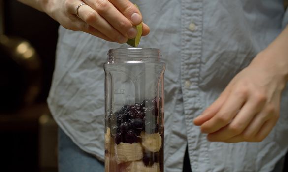 Cooking smoothies in the kitchen. Close up woman's hands squeezing lime juice into the blender glass with banana, blackberry and celery slices. Fresh healthy food