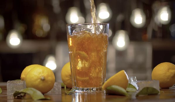 Close up tea pouring into the glass with ice and lemon. Glass with ice tea or cocktail, ice, lemons and leaves on a table against the blurry lights background. Bar counter closeup