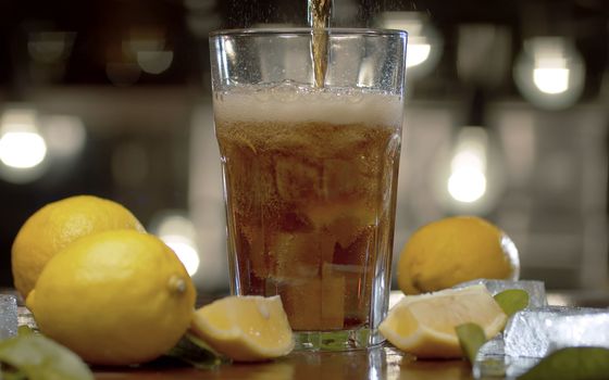 Close up sweet sparkling water pouring into the glass. Fizzy drink with ice and lemon in glass on blurry lights background. Gas escaping from a glass, droplets are clearly visible in back light.