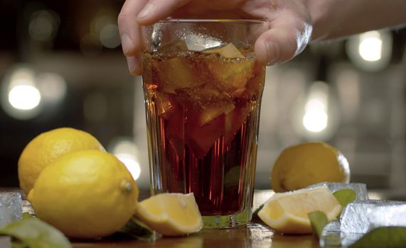 Close up male hand putting a glass on the table. Glass with long island iced tea or cola, ice, lemons and leaves on a table against the blurry lights background. Bar counter closeup