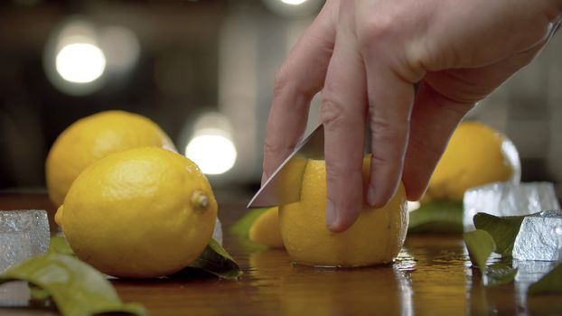 Close up male hand cutting lemon into wedges on the table over background of blurry light bulbs. Cooking of refreshing drinks. Bartender working closeup