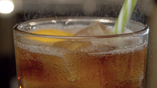 Close up fizzy drink with ice and lemon in glass on blurry lights background. Gas escaping from a glass, droplets are clearly visible in back light.