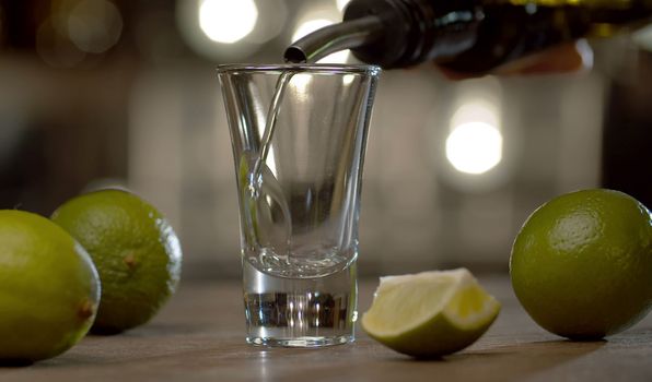 Close up bartender pouring tequila into a glass. Limes on the table. Bar counter on the background of blurry light bulbs.