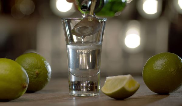 Making a short drink red dog. Close up bartender pouring sambuca into a glass with tequila. Limes on the table. Bar counter on the background of blurry light bulbs.