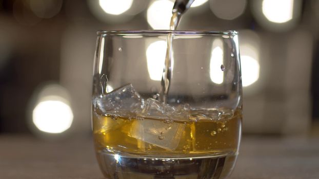 Close up pouring whiskey into drinking glass with ice cubes. Bar counter on the background of blurry light bulbs.