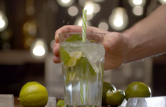 Male hand putting a glass with a tonic, soda on the table. Close up glass with sparkling water, mint, lime and ice over background of blurry lights. Refreshing fizzy drink, mineral water, cocktail.