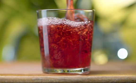 Red transparent juice or wine pouring into the glass with splashes and bubbles against natural green background. Close up cherry or pomegranate or cranberry juice or wine. Healthy eating concept.