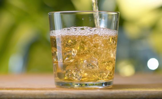 Yellow light transparent juice pouring into the glass with splashes against natural green background. Close up apple, grape juice with ice. Healthy eating concept.