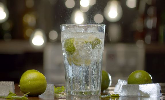 Close up tonic, soda water in a glass with lime and ice on the table. Fizzy refreshing drink, mineral water, cocktail. Gas coming of, jumping water droplets are shining in back light.