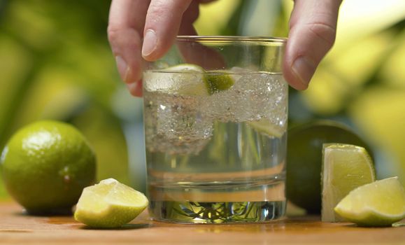 Close up hand shaking a glass of soda water. Tonic fizzy water with ice and piece of lime on a green background. Refreshing mineral water, preparing of drinks, cocktails. Cold lemonade drink