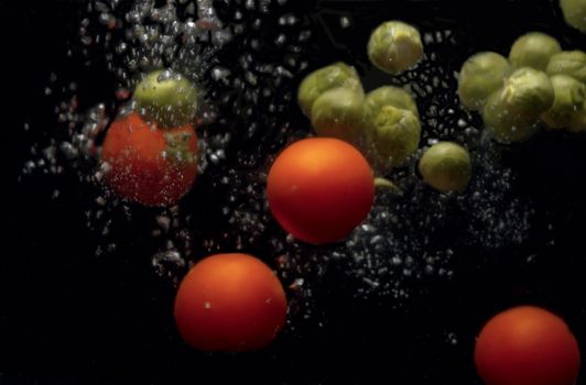 Close up brussels sprouts and tomatoes falling into the water with a splash and bubbles on black background