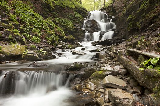 The clear water of a mountain river passes through a cascade of boulders on a waterfall between the hills of the Carpathian Mountains.