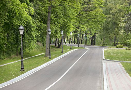 The winding asphalt road goes down the slope through the park, the pavement for pedestrians is paved with stone, a number of beautiful vintage lamps ready to light the way