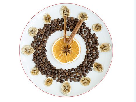 Grains of roasted coffee, mugs of dried orange and banana, cinnamon stick, anise star lie on a porcelain plate in the form of a clock in a bowl isolated on white background