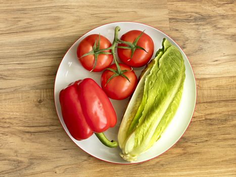 Whole fruits of fresh vegetables close-up: Bulgarian pepper, tomato branch and romaine salad lie on white round porcelain plate on the background of a wooden table