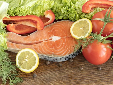 Raw salmon steak on a wooden board. Leaves of lettuce and dill, a tomato branch, spices, slices of lemon and bell pepper, peas of fragrant pepper on a wooden board.