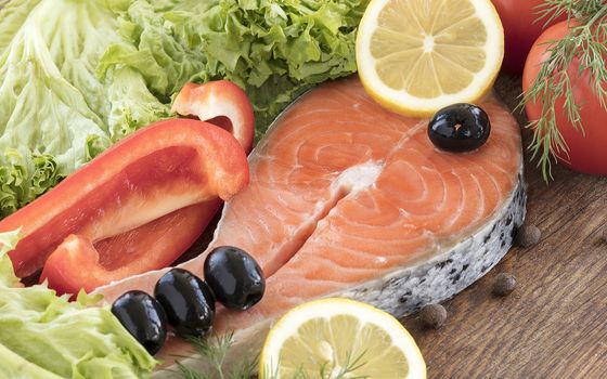 Raw salmon steak on a wooden board. Leaves of lettuce and dill, a tomato branch, black olives, spices, slices of lemon and bell pepper, peas of fragrant pepper on a wooden board.