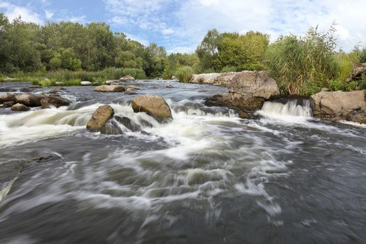 The rapid flow of the river, rocky shores, rapids, bright green vegetation and a cloudy blue sky in the summer. Southern Bug. Ukraine