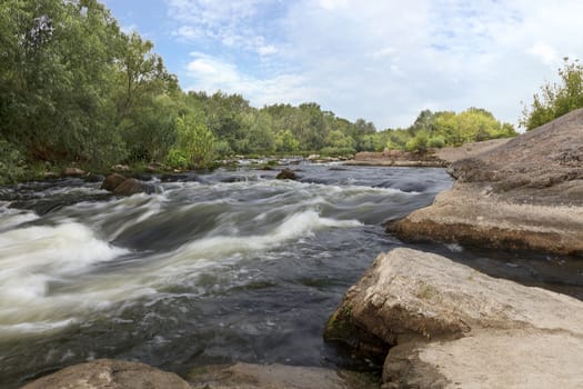 Rocky shores, rapids, fast river flow, bright green vegetation and a cloudy blue sky in summer