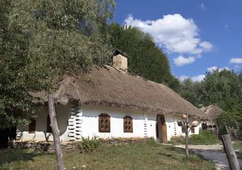 Old traditional Ukrainian rural house with thatched roof and wicker fence in the garden against a blue sky with a white cloud