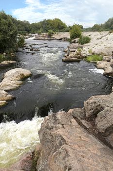 A view of the river rapids and the rapid water flow of the Southern Bug River in the summer. A teenager stands on the edge of a stone boulder on the river bank.