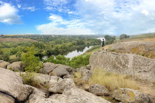 The teenager stands on top of a large stone boulder on the bank of the South Bug River and looks at the river below. The river Southern Bug in the summer - river flow, rocky shores, bright green vegetation and a cloudy blue sky