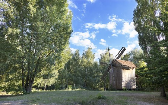 An old wooden windmill on a green glade in a summer young forest, against a blue sky with light white clouds