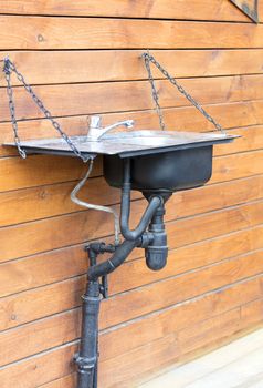 A modern washbasin sink with a stainless steel faucet installed outside on a chain on the wall of a wooden house, in a rustic farmhouse