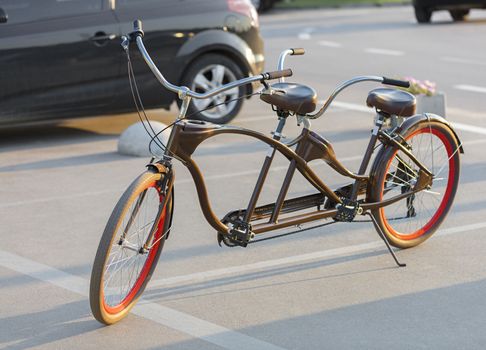 A chocolate-colored tandem bicycle with scarlet wheels is parked in a car park near the bike path in the evening sun.