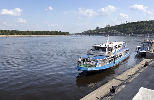 Pleasure boat moored at the city wharf of the Dnieper River in Kiev against the background of a bridge across the river and urban wooded park slopes
