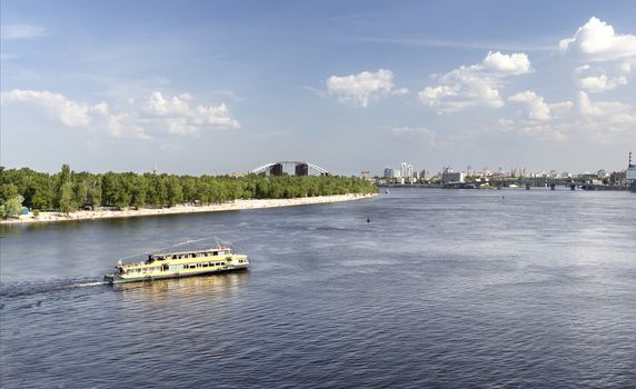 A pleasure boat walks along the Dnieper River on a bright summer day against the backdrop of the city beach, the blue sky and the city skyline of Kyiv