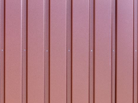 Reddish-brown metal fence made of corrugated steel sheet with vertical guides and metal rivets in the middle. Corrugated reddish-brown iron sheet background close up.