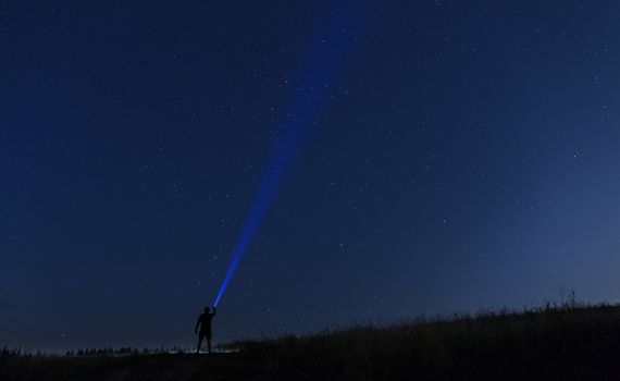 Silhouette of man on a background of stars he directs a ray of light onto a dark blue starry sky