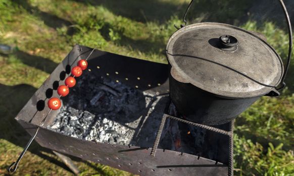 In the old cauldron on the barbecue cooking porridge against a forest clearing at noon. On the skewer are strung and baked red tomatoes.