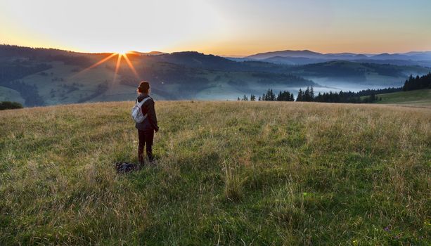 A young woman in the rays of the rising sun meets the dawn in the meadow of the hill and looks at the misty morning mountains of the Carpathians