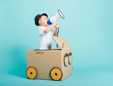 Happy Asian children boy smile in driving play car creative by a cardboard box imagination with megaphone, summer holiday travel concept, studio shot on blue background with copy space for text