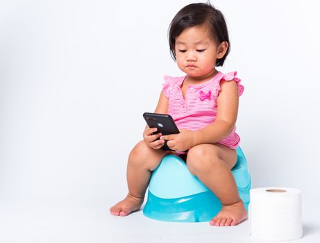 Asian little cute baby child girl education training to sitting on blue chamber pot or potty and play smart mobile phone with toilet paper rolls, studio shot isolated on white background, wc toilet