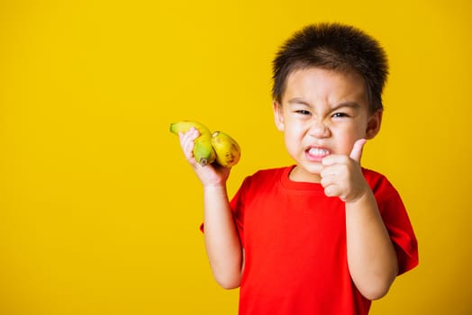 Happy portrait Asian child or kid cute little boy attractive smile wearing red t-shirt playing holds bananas and show finger thumb for good sign, studio shot isolated on yellow background