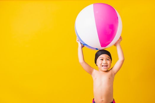 Summer vacation concept, Portrait Asian happy cute little child boy smiling in swimsuit hold beach ball, Kid having fun with inflatable ball in summer vacation, studio shot isolated yellow background