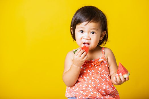 Happy portrait Asian baby or kid cute little girl attractive laugh smile playing holds cut watermelon fresh for eating, studio shot isolated on yellow background, healthy food and summer concept