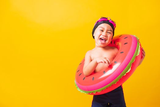 Summer vacation concept, Portrait Asian happy cute little child boy wear goggles and swimsuit hold watermelon inflatable ring, Kid having fun on summer vacation, studio shot isolated yellow background